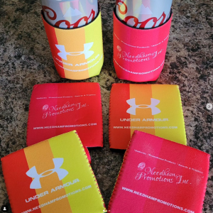 Five orange-to-yellow gradient can and bottle sleeves lie on a granite countertop; two hold drinks. The sleeves are branded with the Under Armour and Needham Promotions Inc. logos in white along with their website, www.needhampromotions.com.