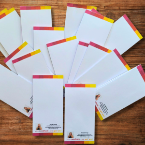 A fan of rectangular notepads with white pages and colorful yellow, orange, and pink borders sits on a wooden surface. The lower left features a headshot of a woman and text reading, 